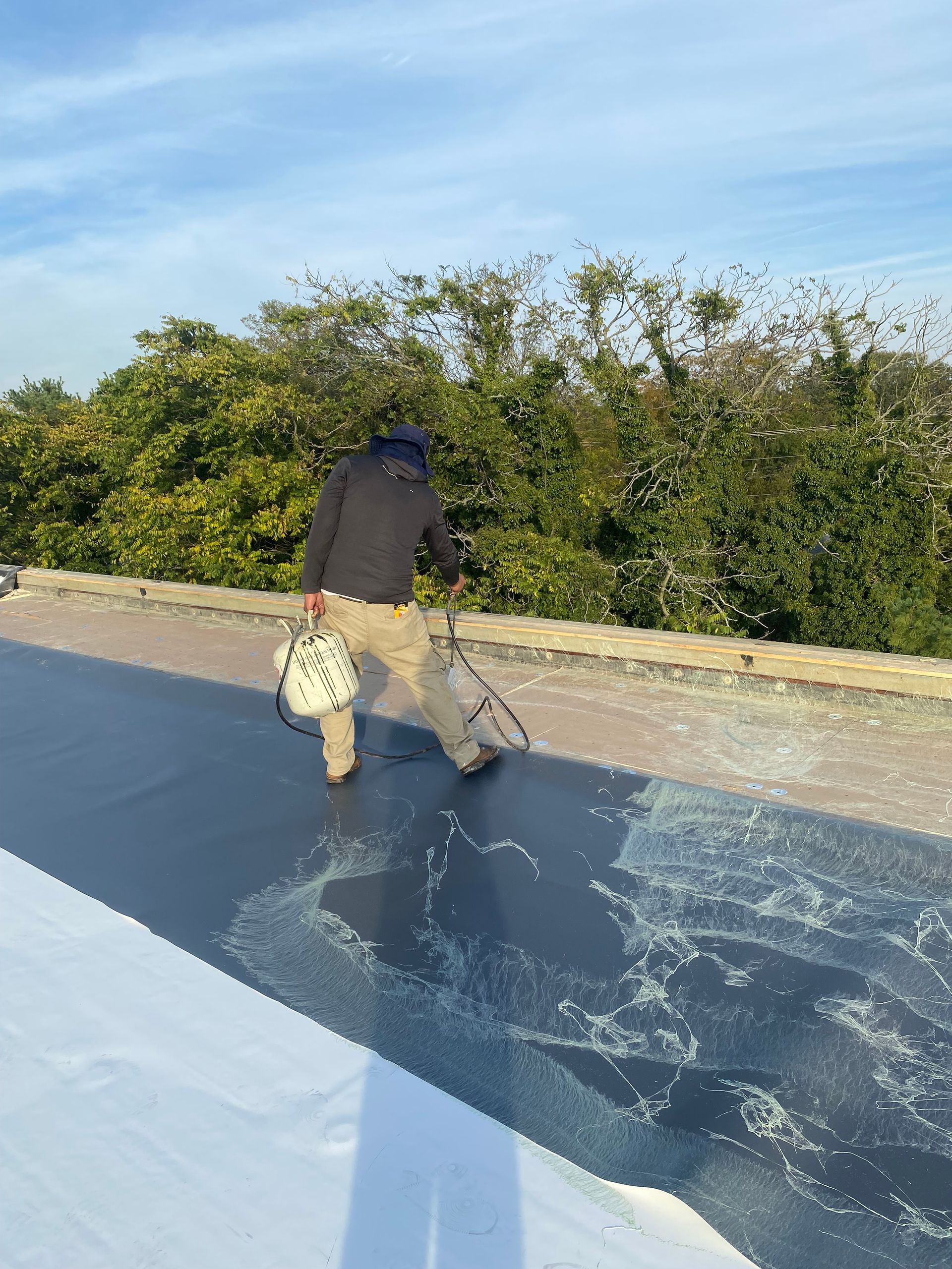 Worker applying dark coating to a flat rooftop beside green bushes under a blue sky