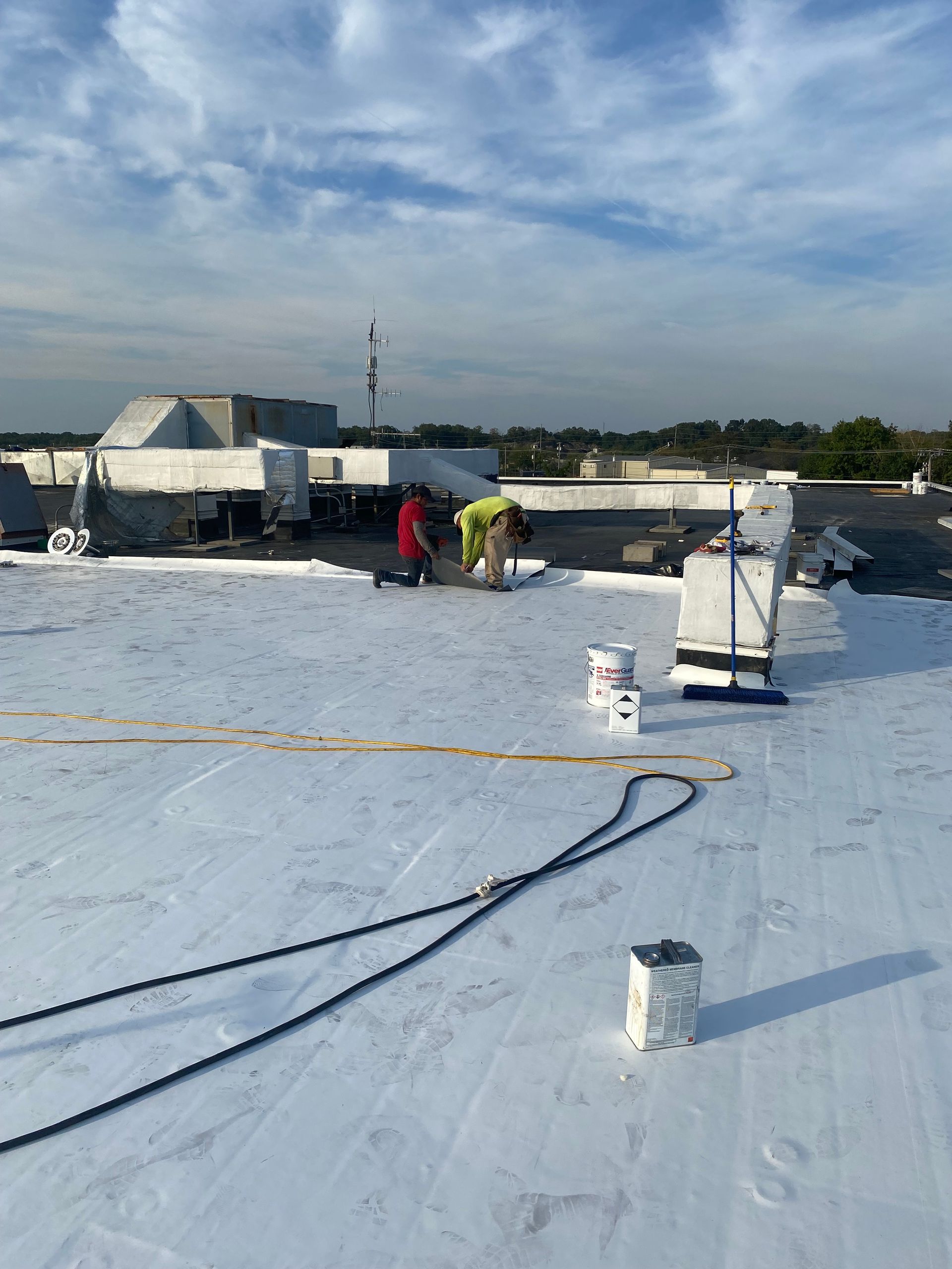 Workers on a flat rooftop installing white roofing material under a cloudy sky.
