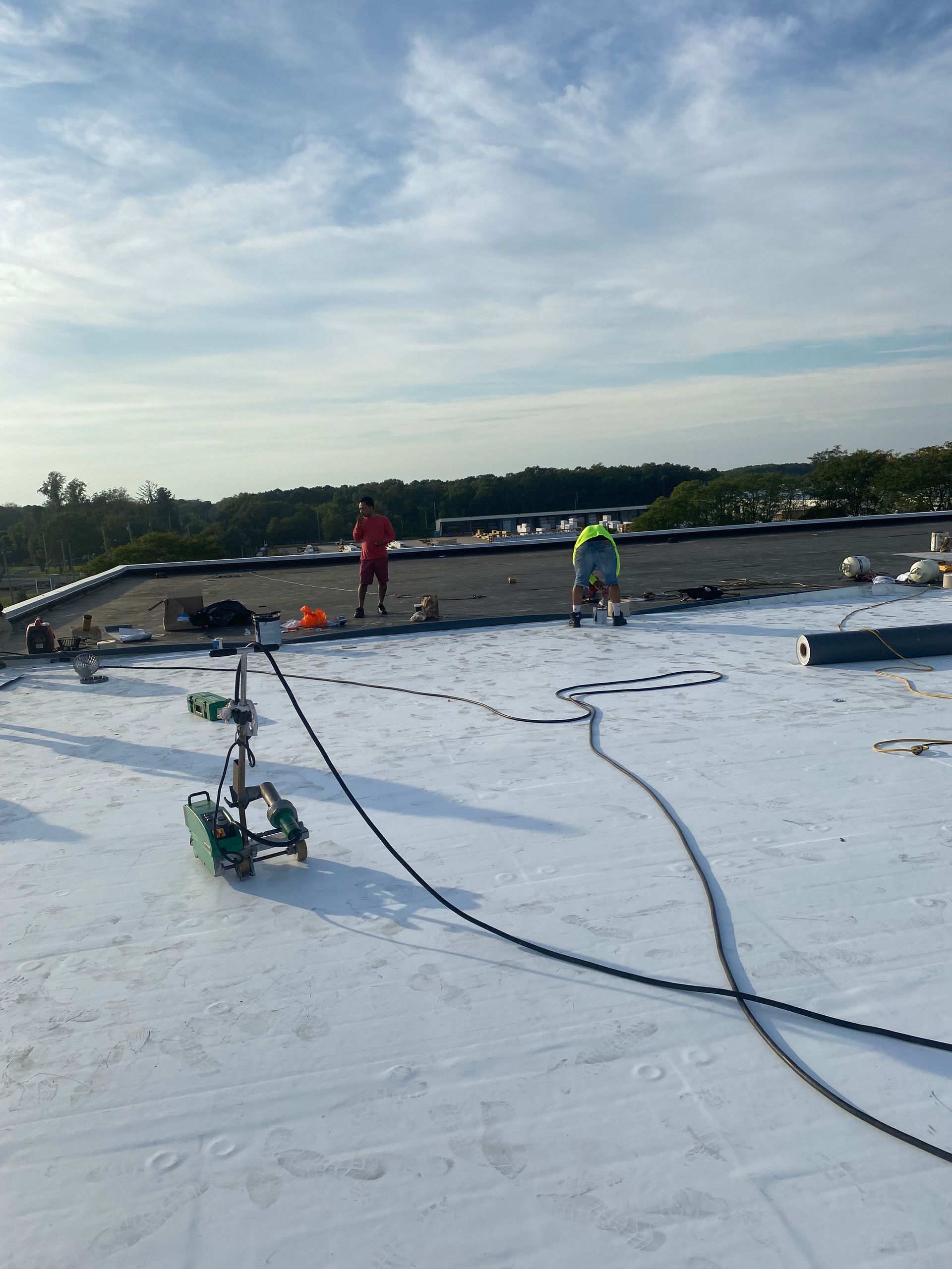 Workers on a white roof securing cables under a cloudy sky, with trees in the background