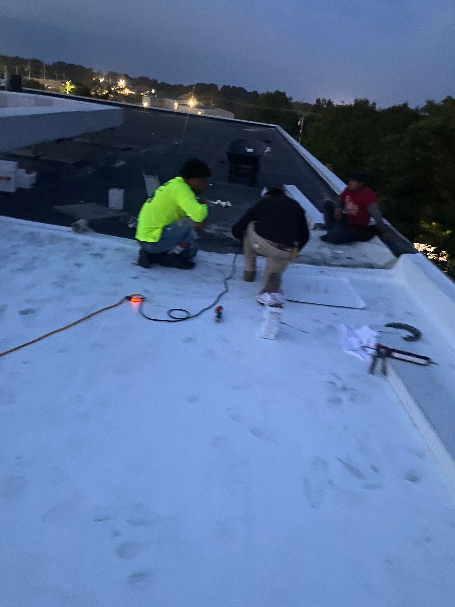 Two workers kneeling on a flat rooftop at dusk, inspecting roofing materials and tools.