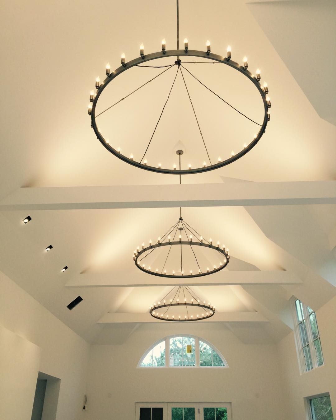 Three circular chandeliers hanging from a white ceiling. The chandeliers are lit and positioned at different heights.