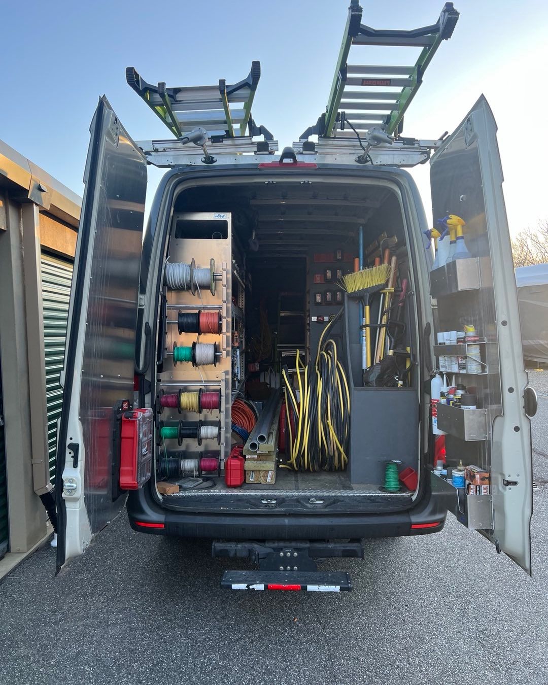 Back of a white work van with doors open, ladder on top, tools and supplies inside, parked outdoors.