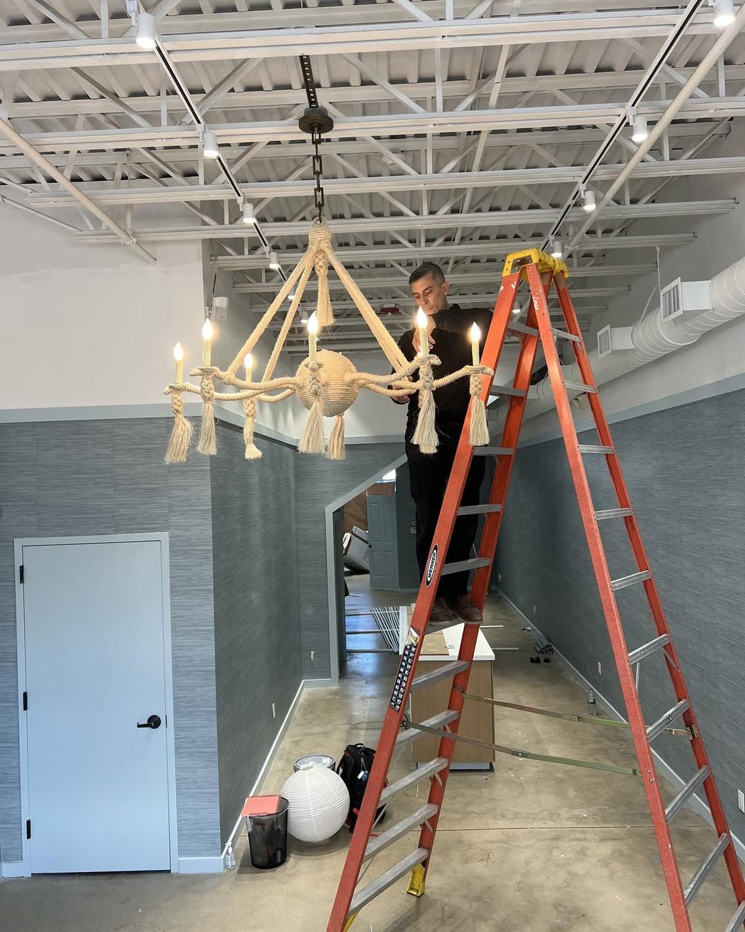 Man on a ladder installing a rope chandelier in a hallway with textured gray walls and a white ceiling.
