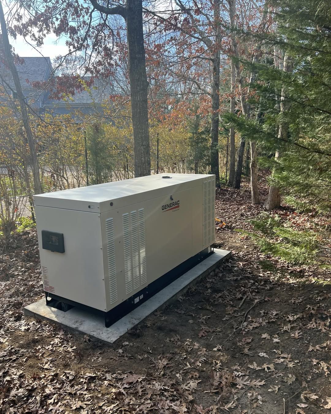 Generator on a concrete pad in a wooded area, surrounded by trees and fallen leaves.