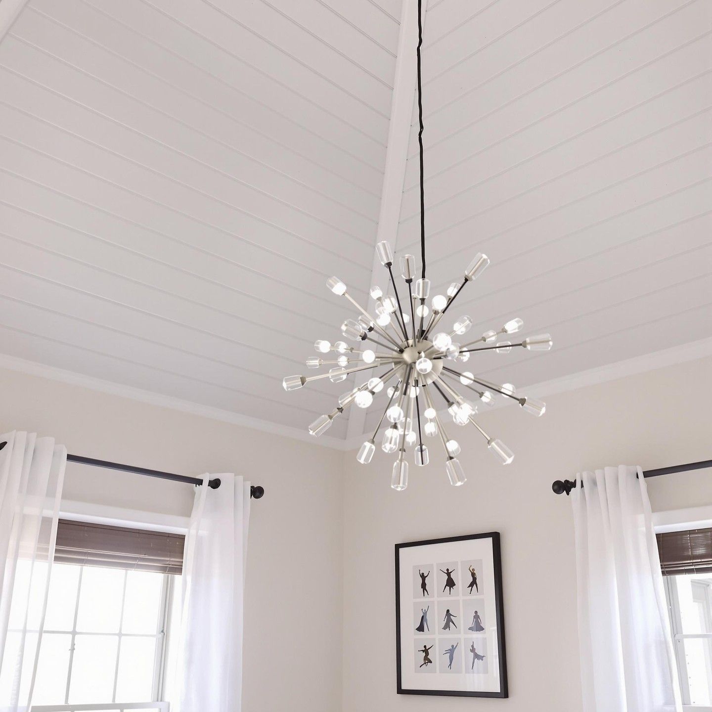 Modern chandelier hangs from a white ceiling. Two windows with sheer curtains and a framed artwork below.