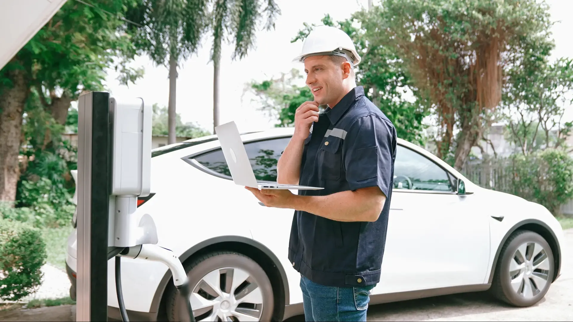 Man in hard hat using a laptop next to an EV charging station and a white electric car.