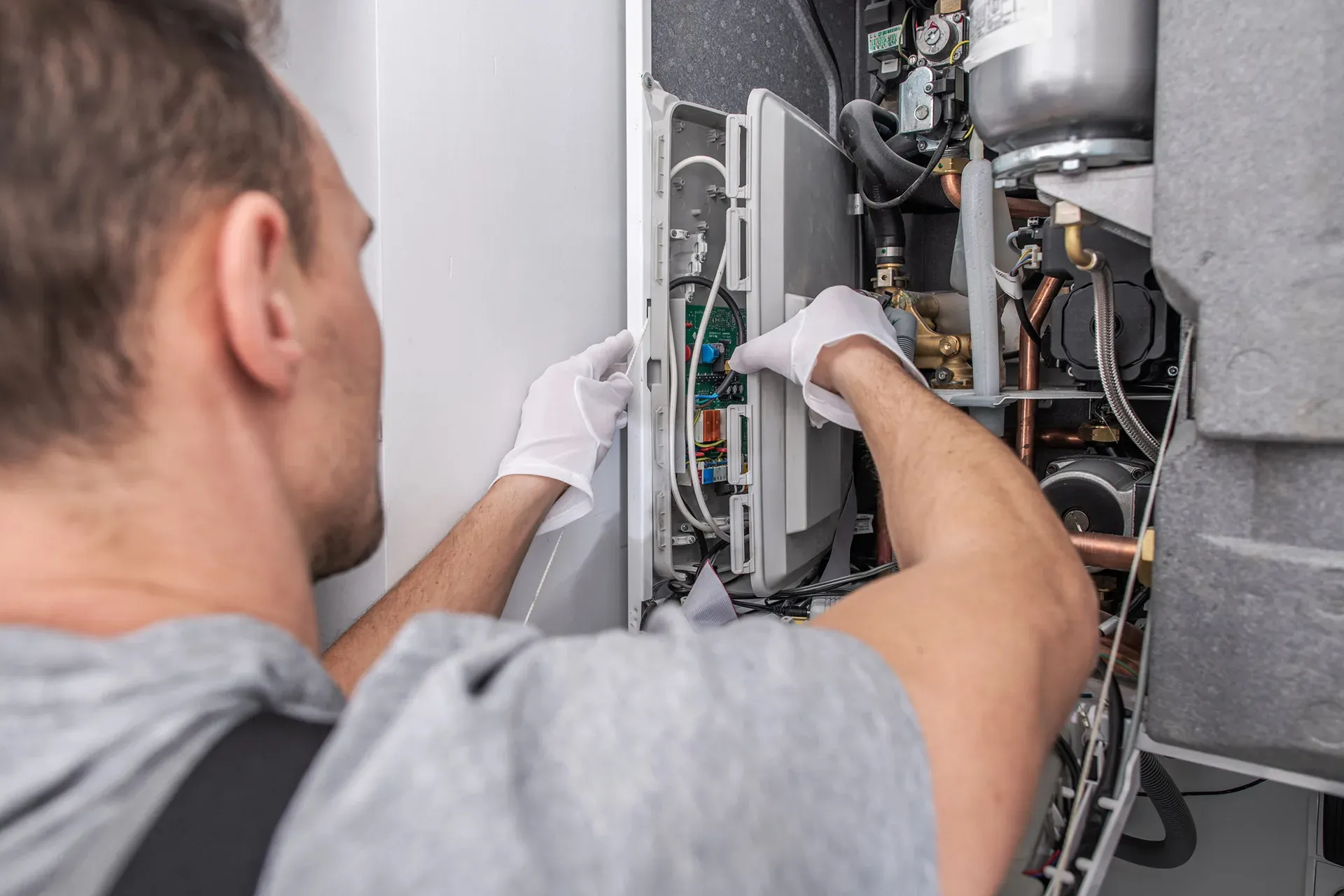 Man in gloves repairing a boiler, hands working near wires and pipes.