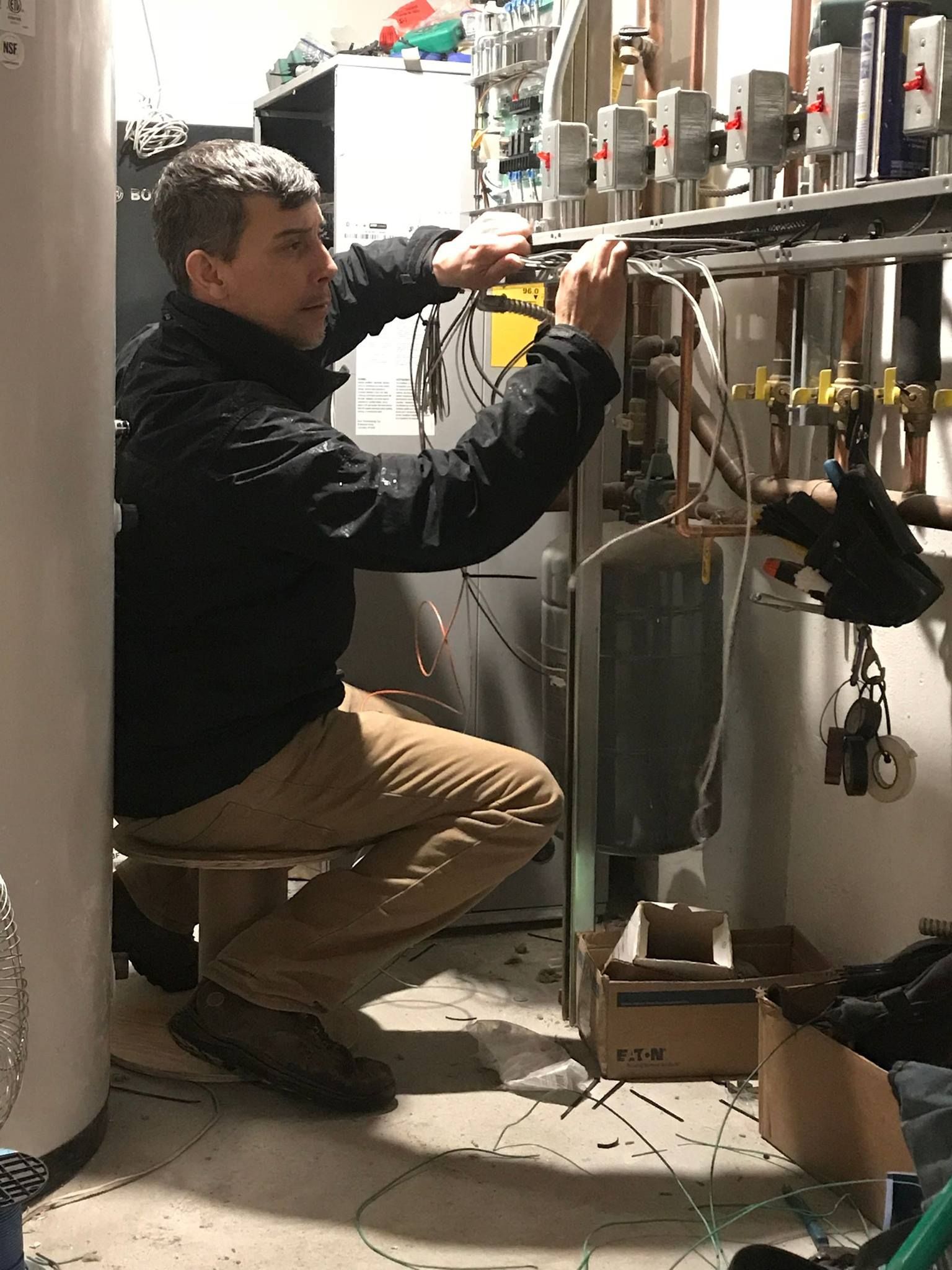 Man in black jacket works on pipes near a boiler in a basement.
