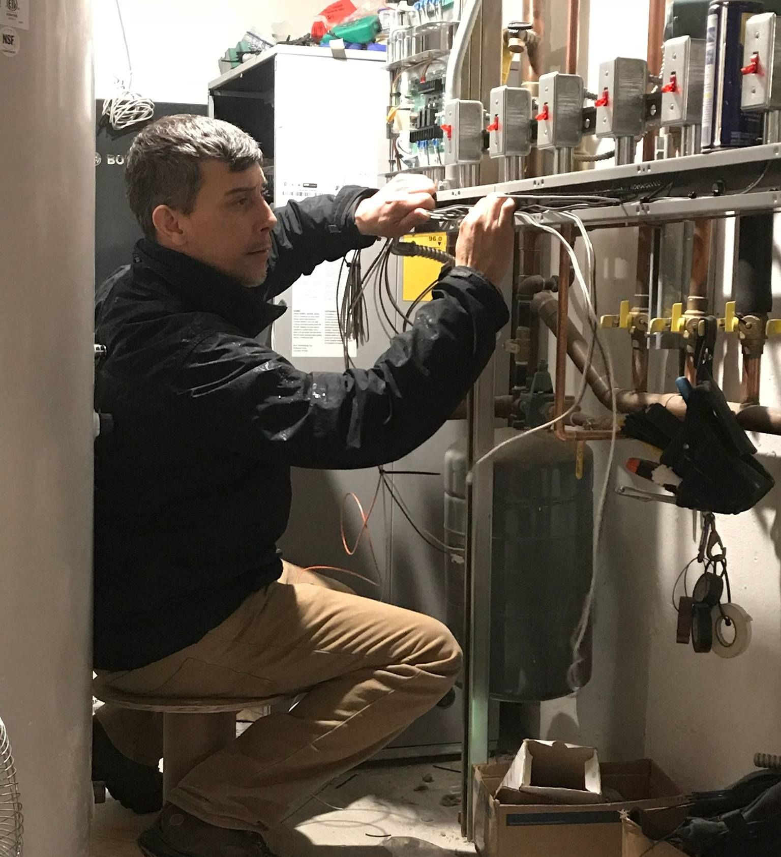 Man in black jacket works on pipes in a utility room, sitting on a stool.