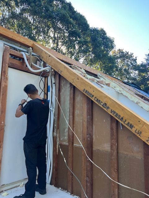 A person installing electrical wiring on a building's exterior wall. Sunlight, trees in the background.