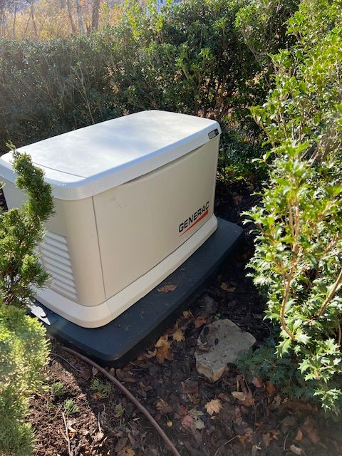 A tan Generac generator on a black base, surrounded by greenery in an outdoor setting.