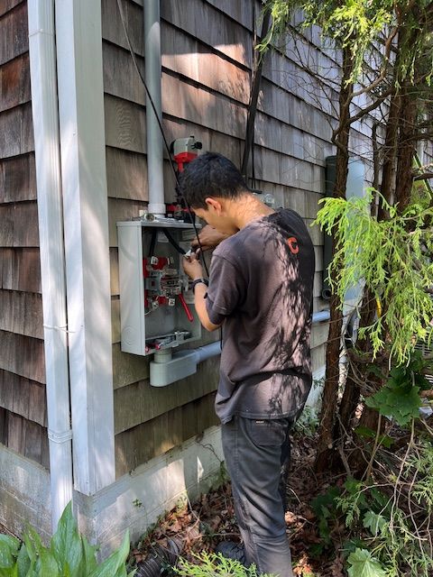 A person working on an electrical box mounted on the side of a shingled building, outdoors.