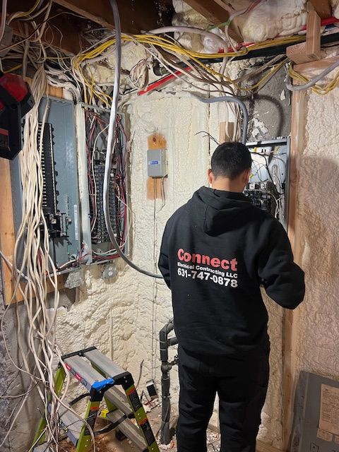 An electrician in a black hoodie works on a circuit breaker panel in a basement with exposed wiring.