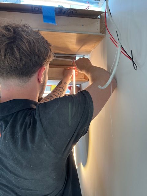 Person installing LED strip lights under a wooden shelf. Wires and electrical work visible.