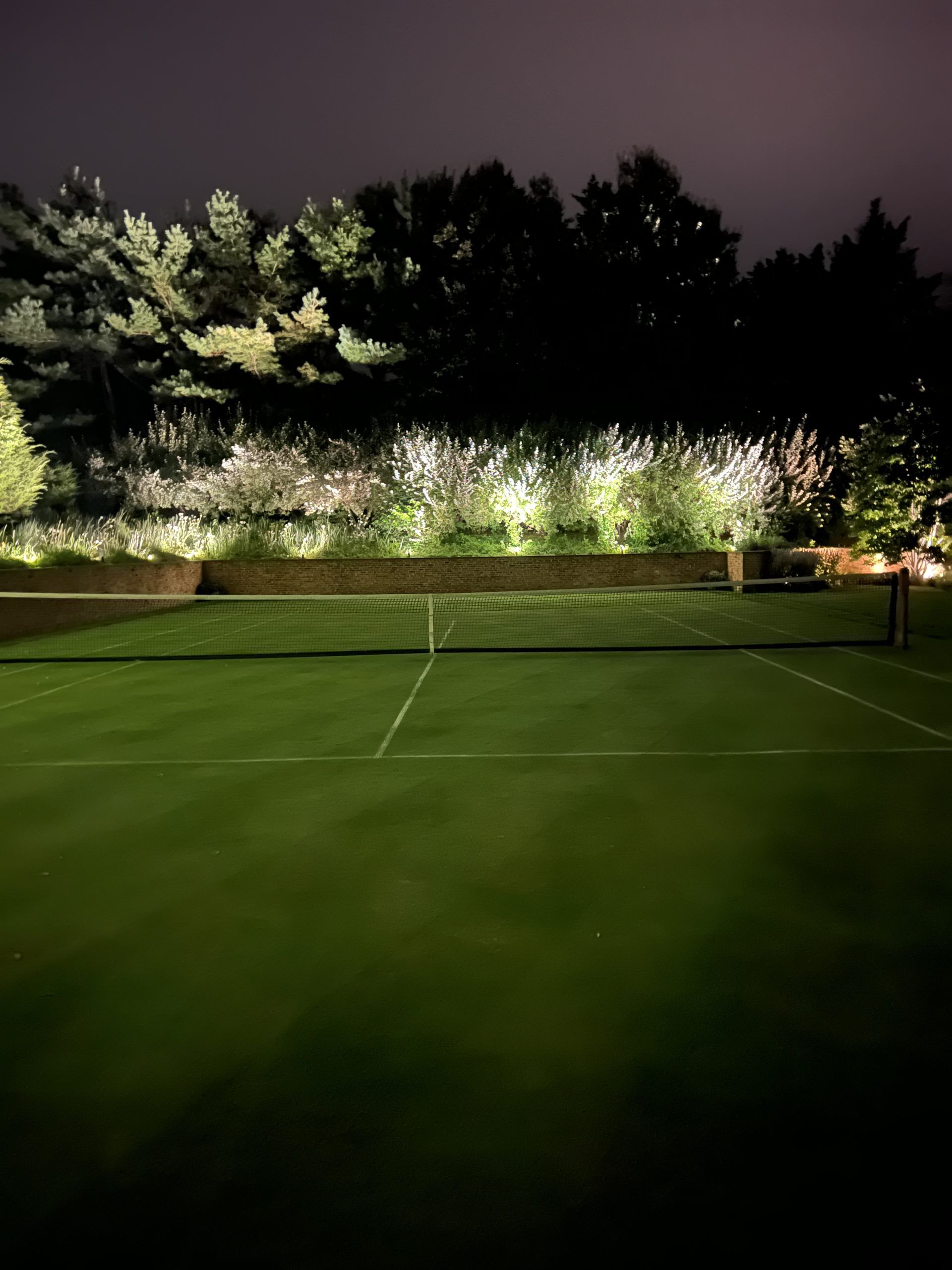 Lit tennis court at night, green surface, net, illuminated trees in the background.