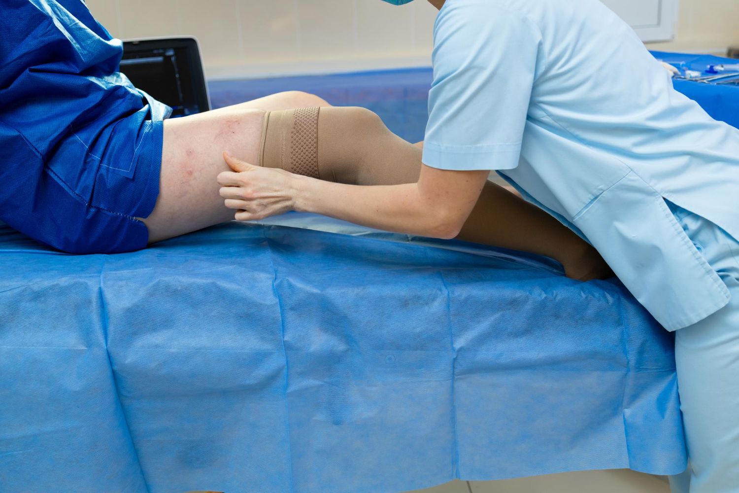 Healthcare worker examining a patient’s leg on an examination table, with blue drapes and gown.