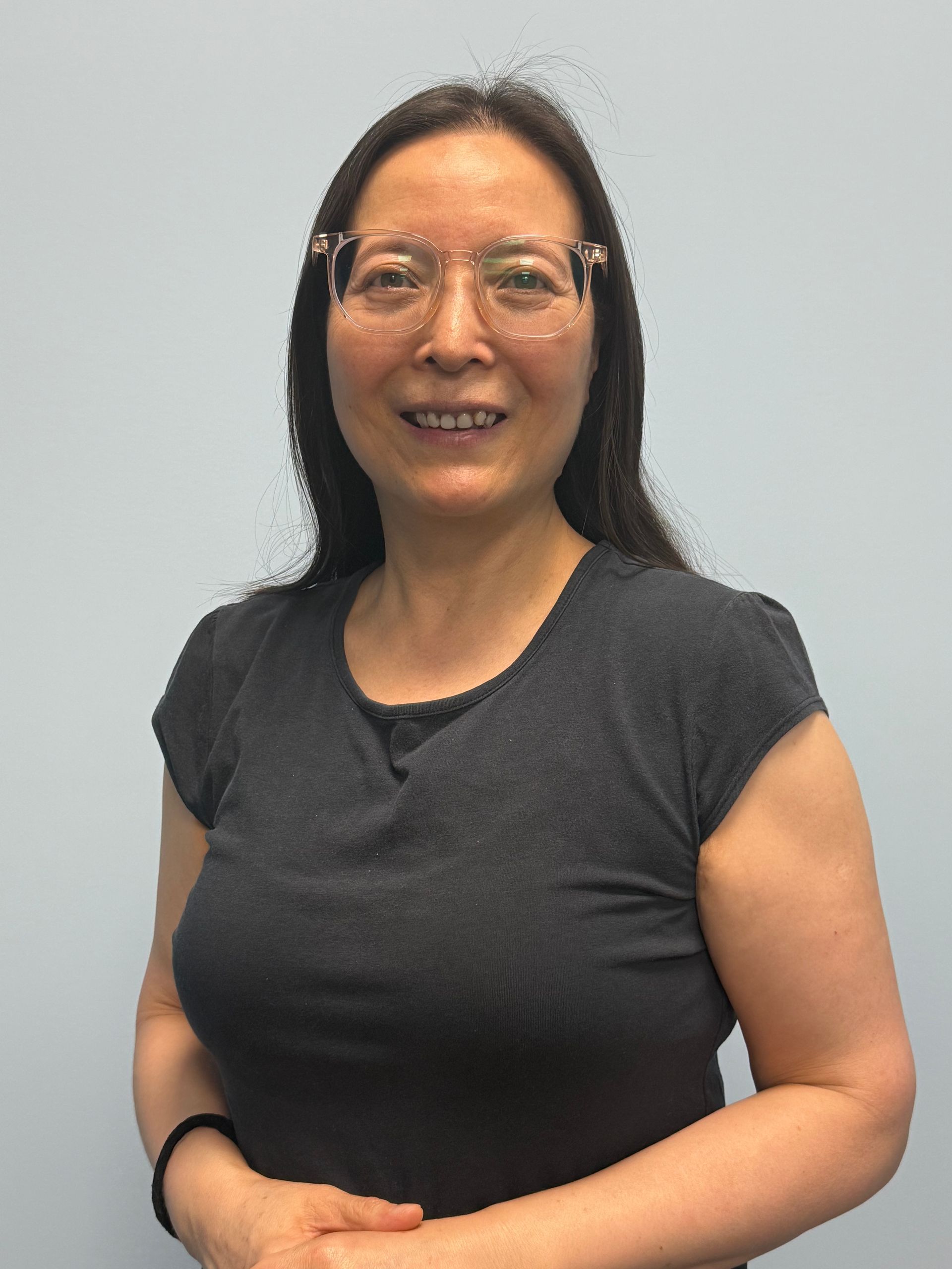 Woman wearing glasses and a dark shirt, smiling while standing against a plain light background.