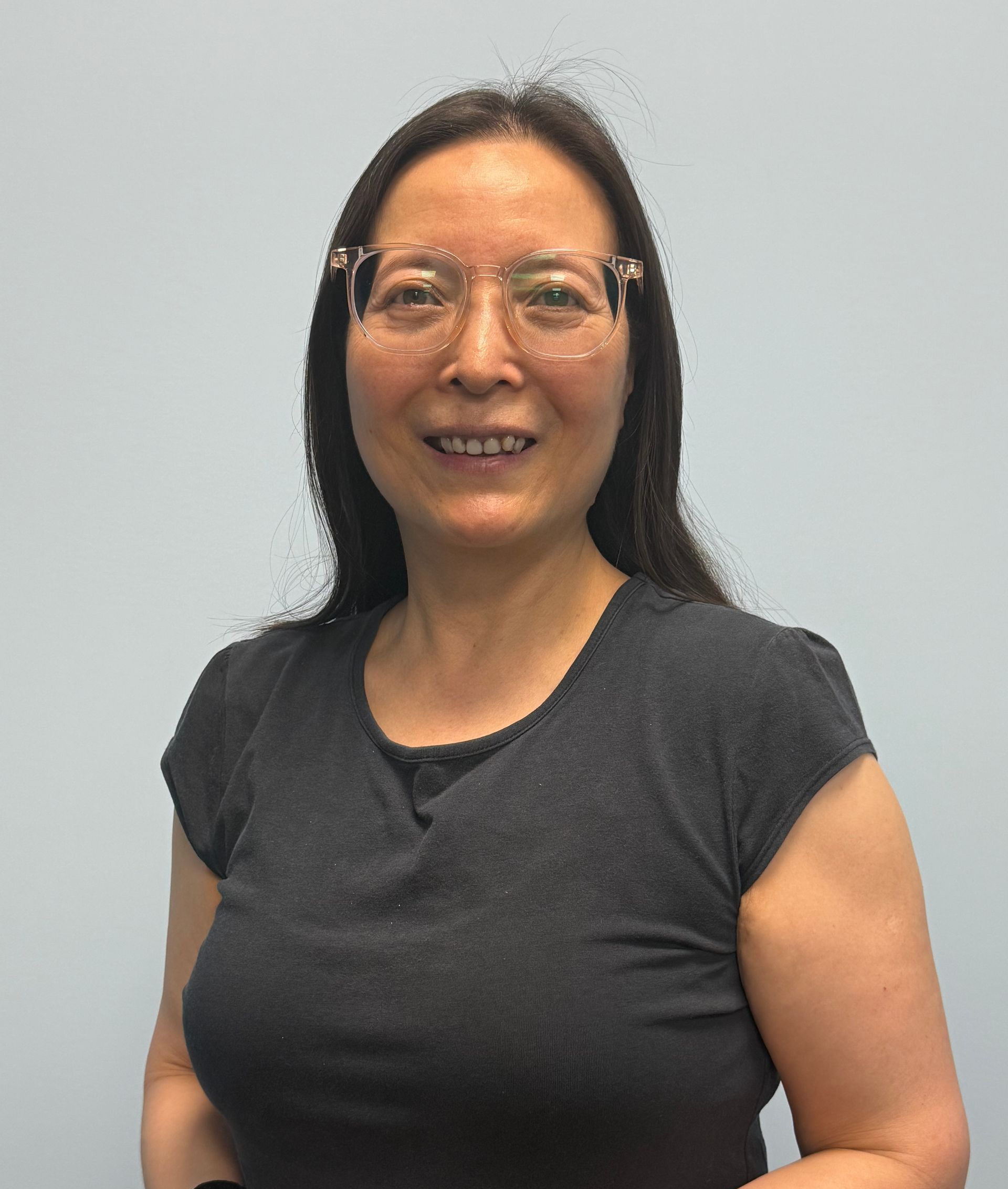 Woman with glasses smiles, wearing a dark gray shirt. Pale gray background.