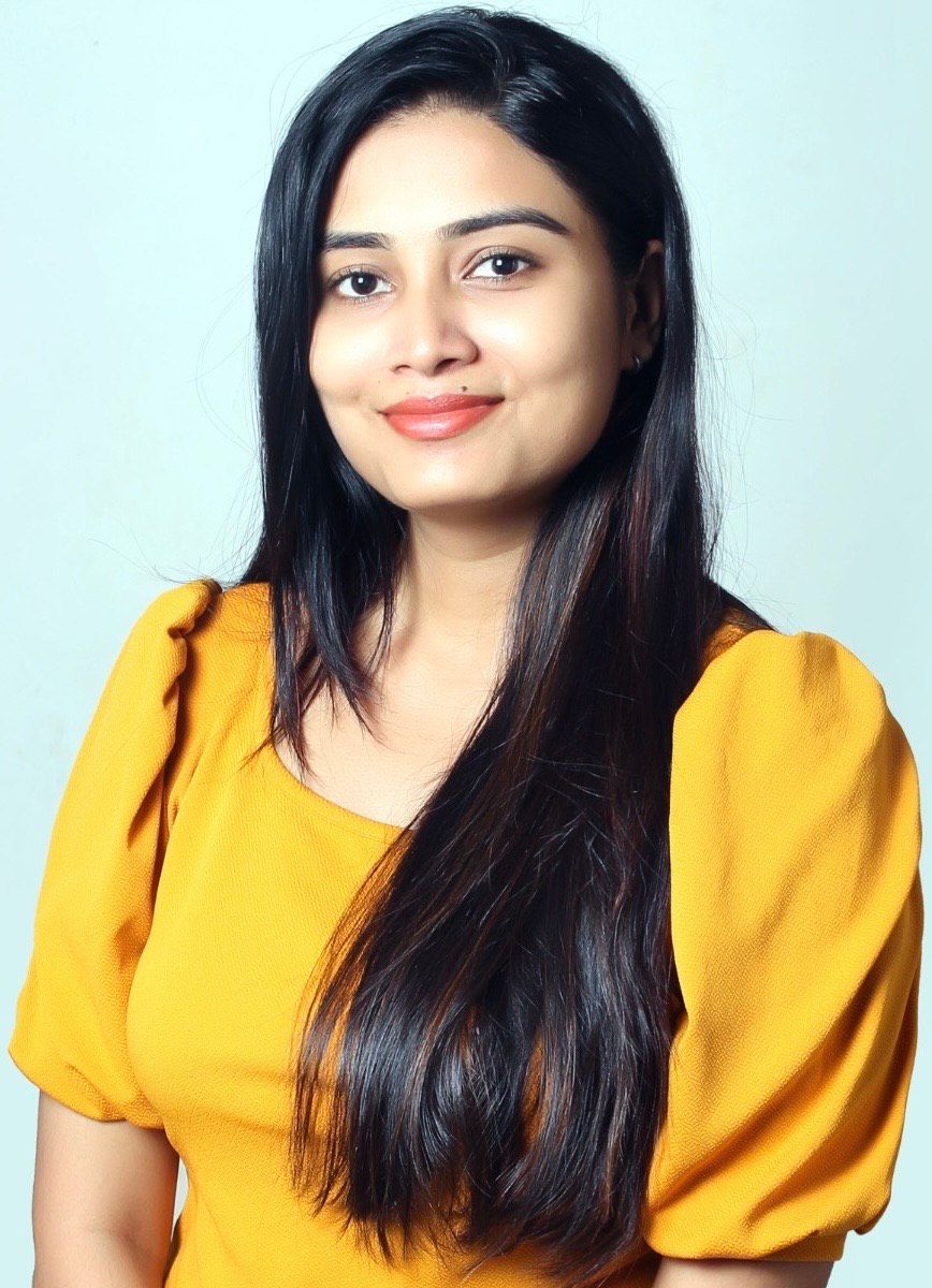 Woman with long black hair wearing a mustard yellow top, smiling against a light blue background