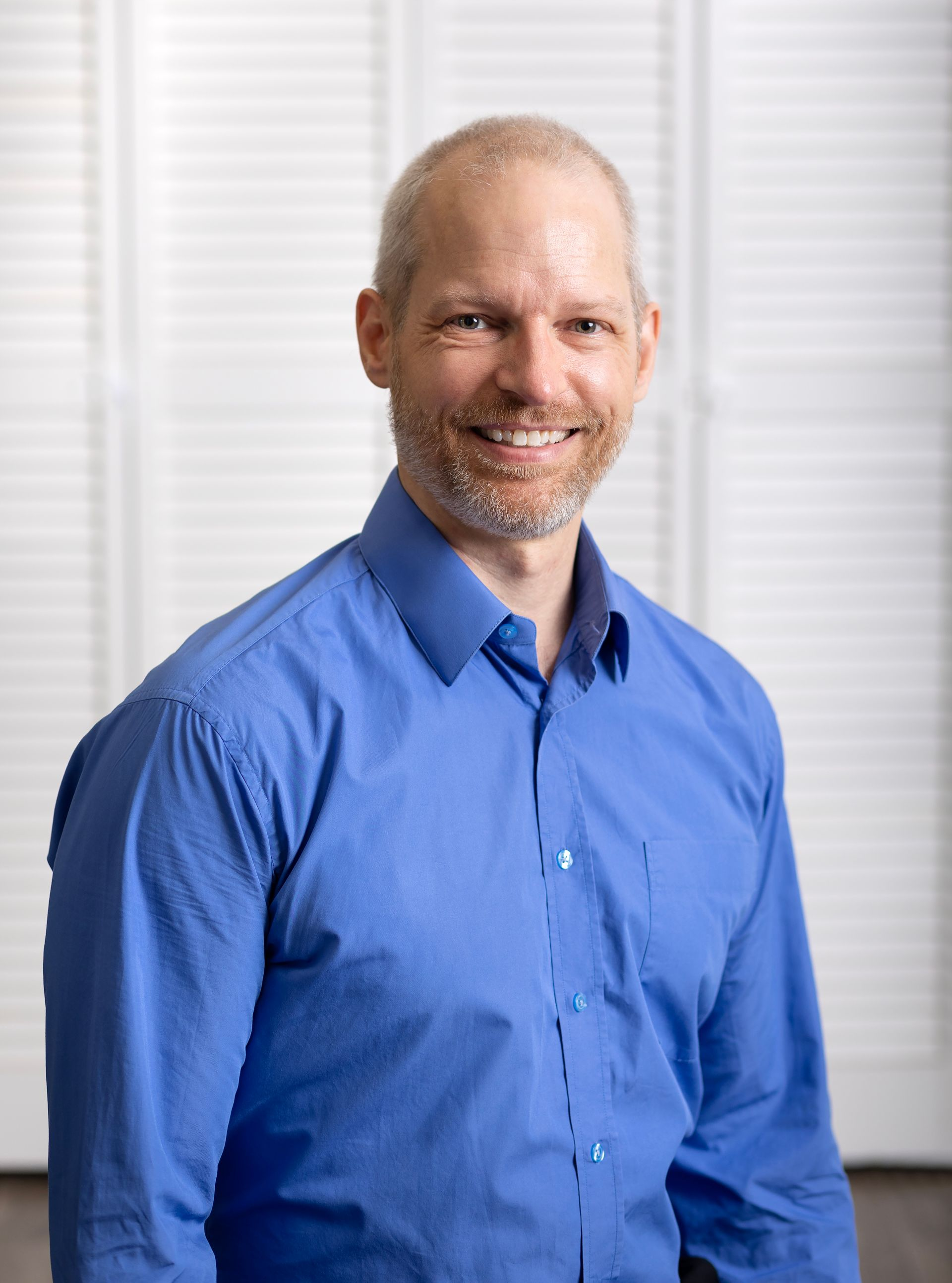 Man in a blue shirt smiling in a bright indoor setting with white blinds