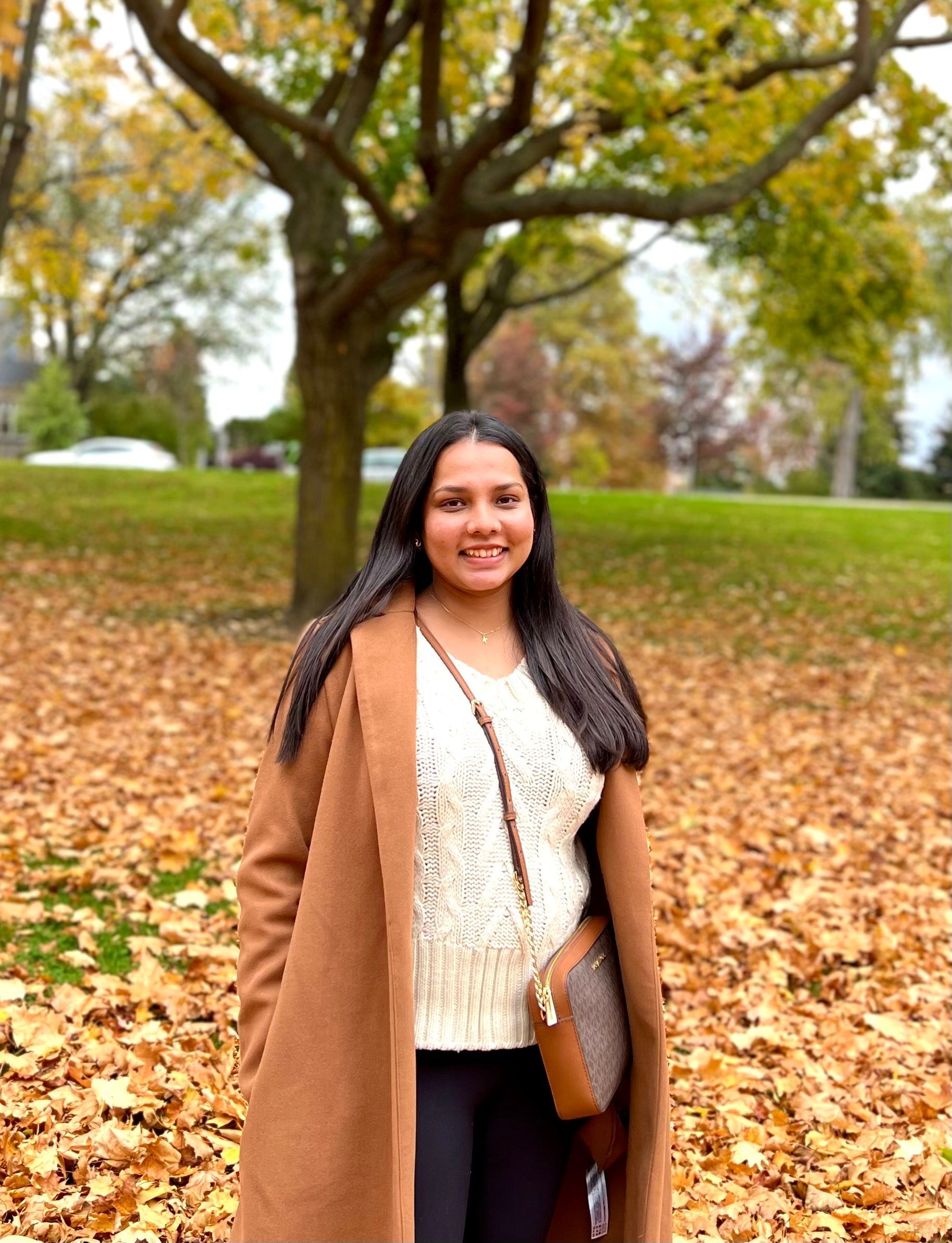 Woman standing in a park covered with autumn leaves, smiling and wearing a tan coat.