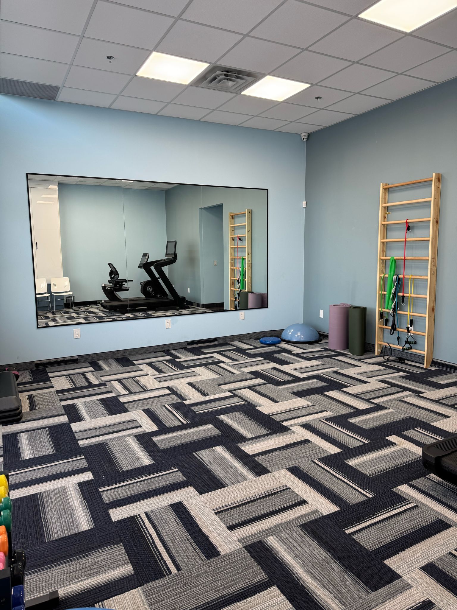 Empty exercise room with patterned carpet, wall mirror, ceiling lights, and yoga equipment against blue walls