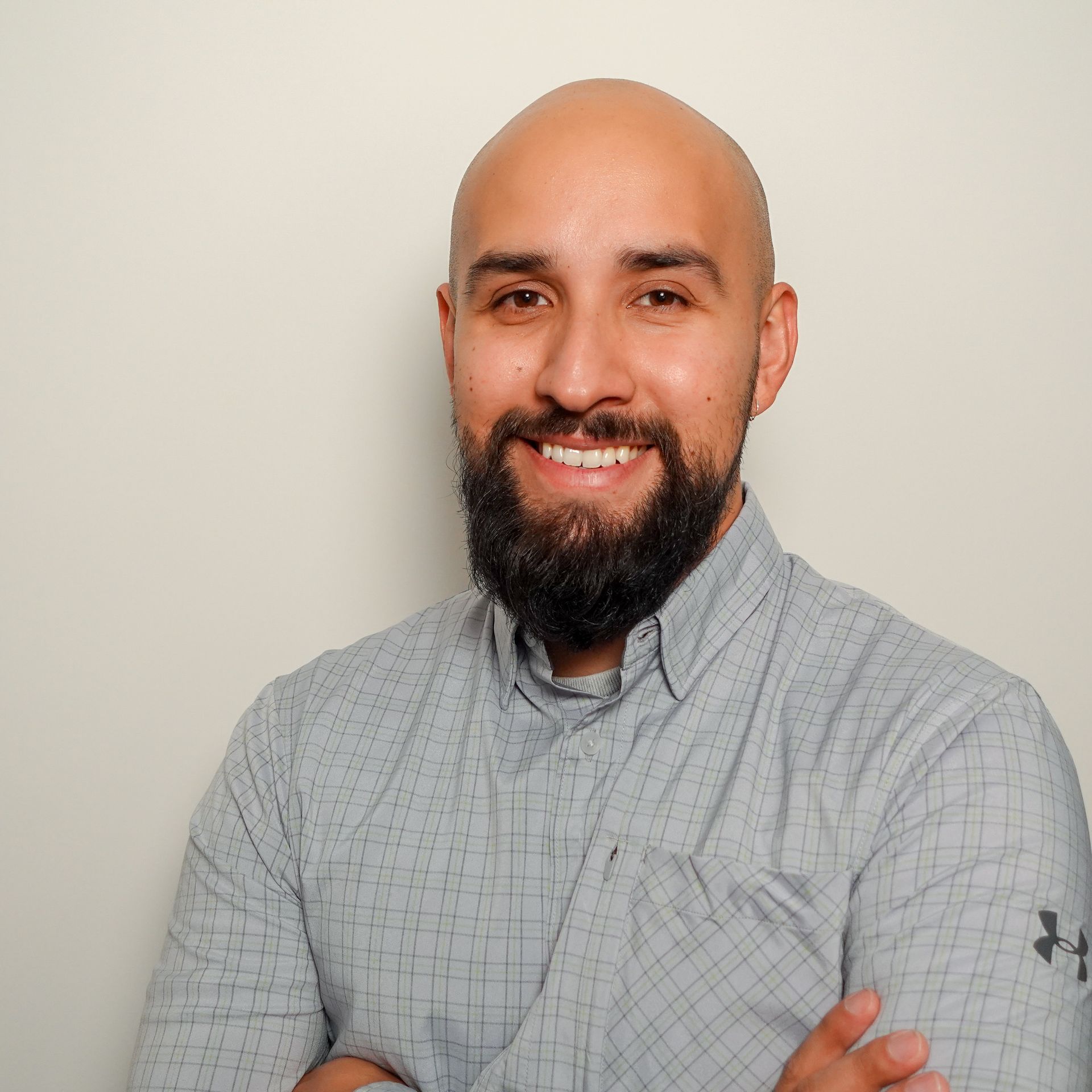 Smiling bald man with a dark beard and crossed arms, wearing a gray button-up shirt against a plain wall