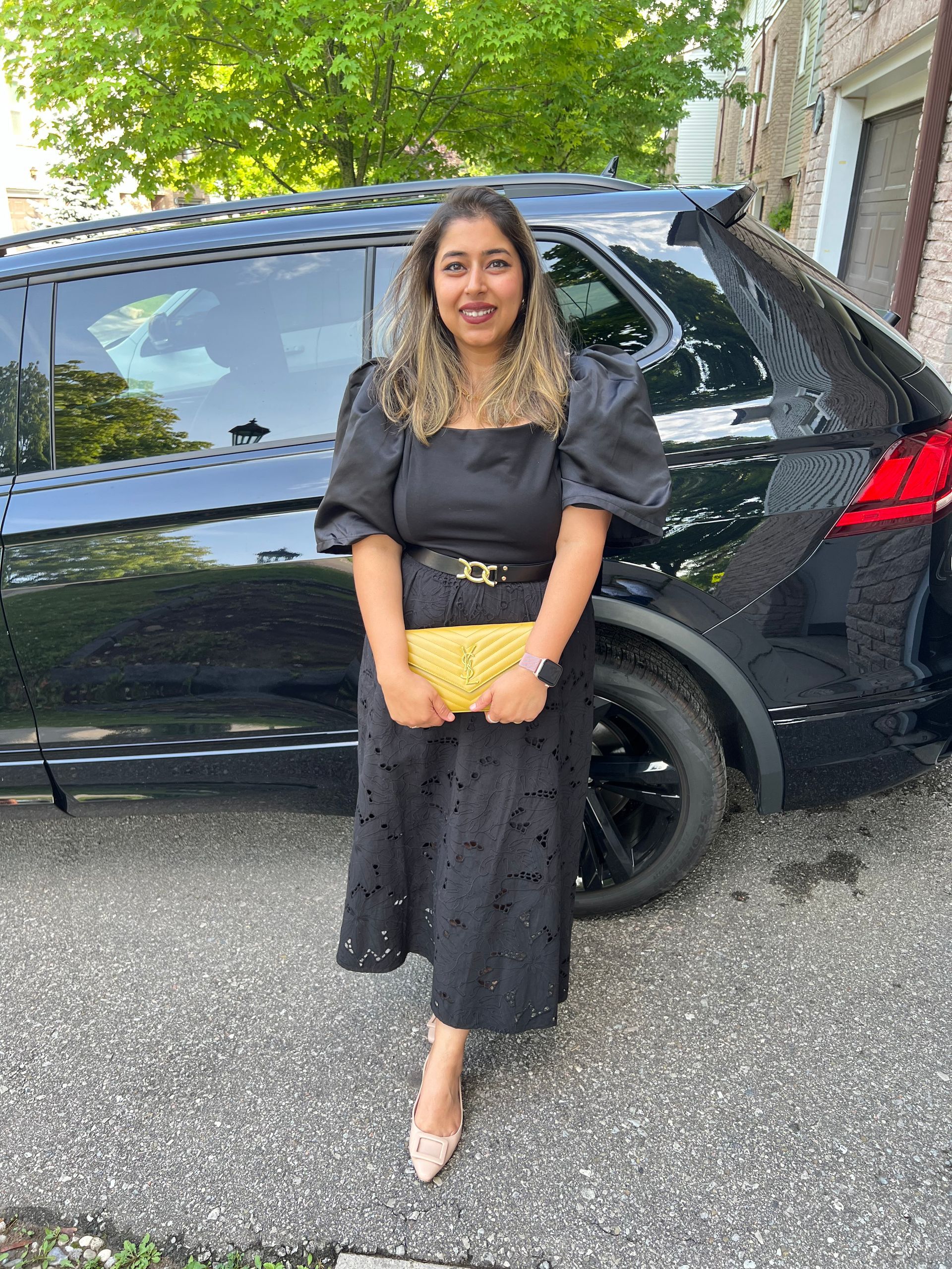 Woman in a black dress stands beside a black SUV, holding a yellow clutch outdoors.