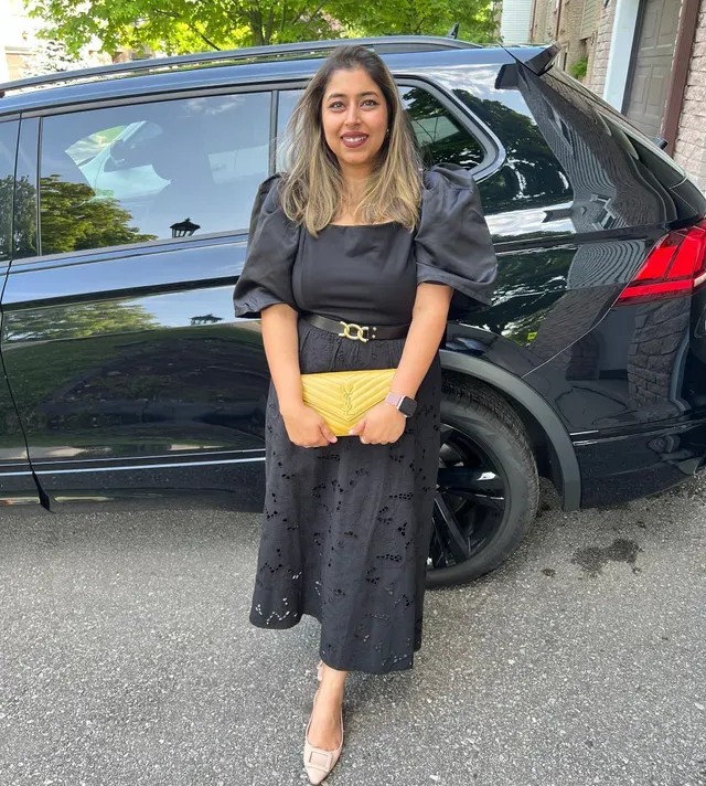 Woman in a black dress stands beside a black SUV, holding a yellow clutch outdoors.