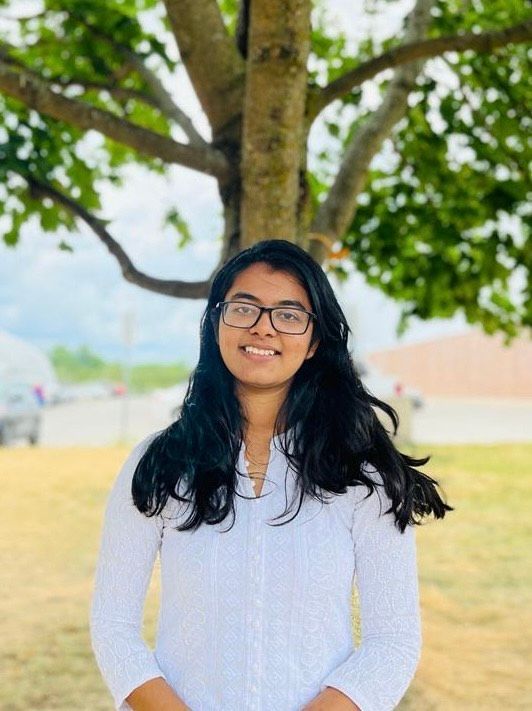 Person smiling in glasses, standing outdoors under a tree in a white shirt.
