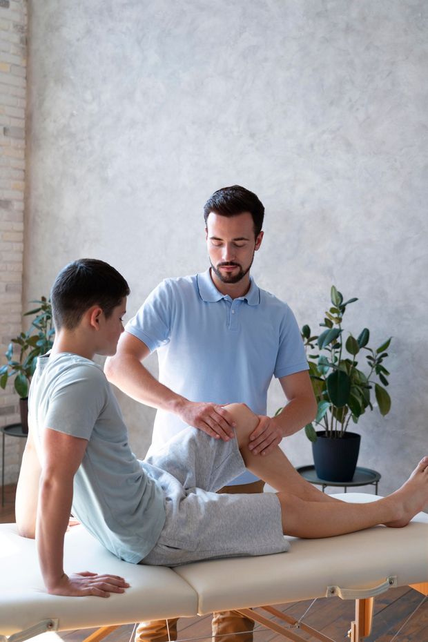 Physiotherapist examining a patient's knee on a massage table. Both are indoors.