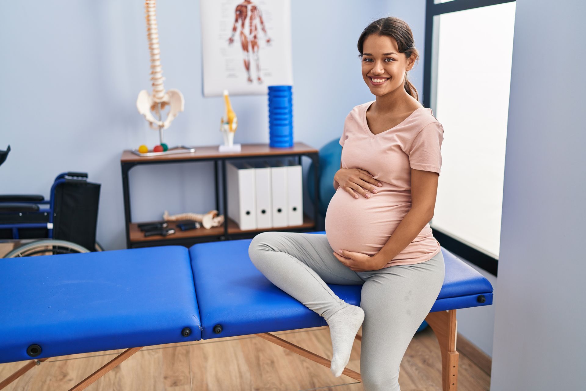 A woman is sitting on a pilates ball while a female physical therapist holds dumbbells.