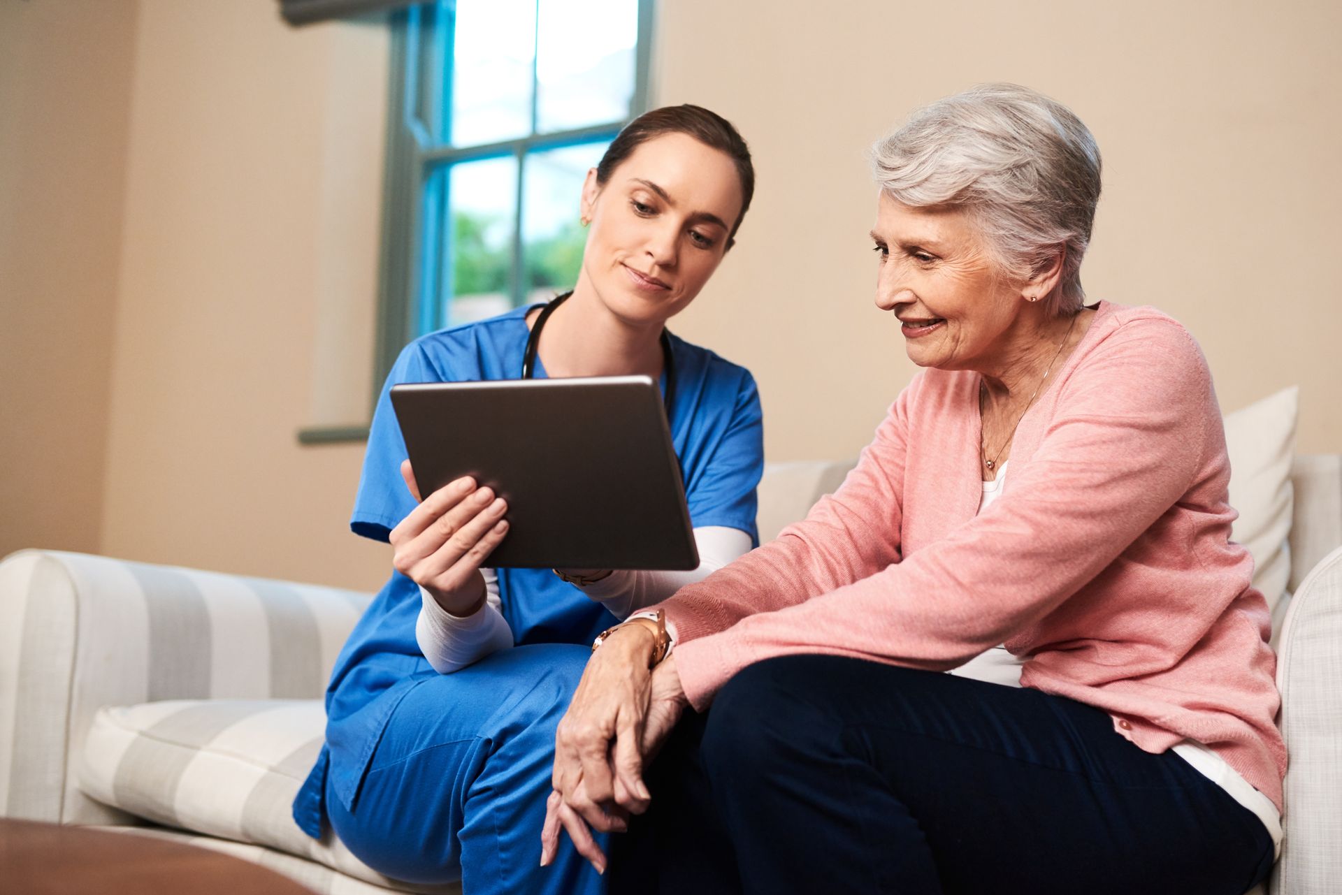 Nurse in blue scrubs shows a tablet to a smiling senior woman sitting on a couch.