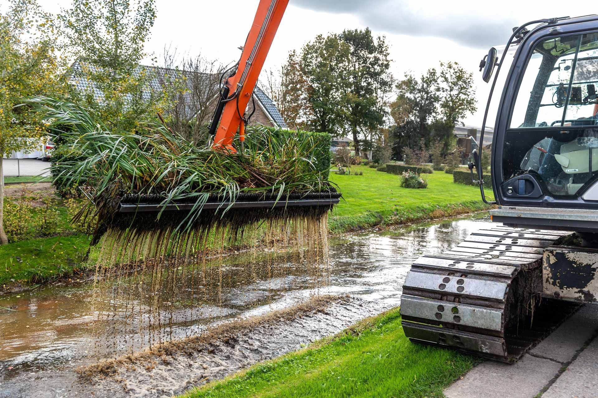 Excavator clearing a flooded roadside ditch beside a grassy field and trees