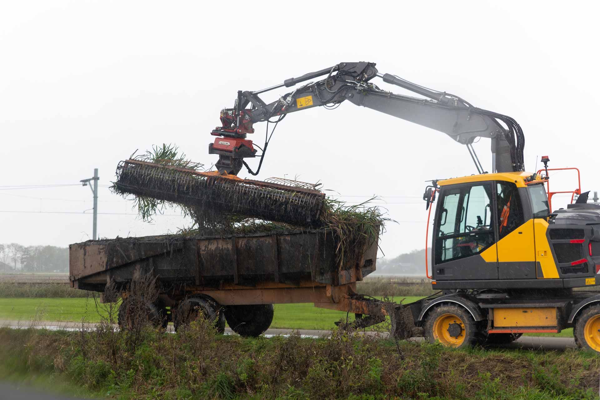 Excavator loading branches into a truck in a rural field under overcast skies.