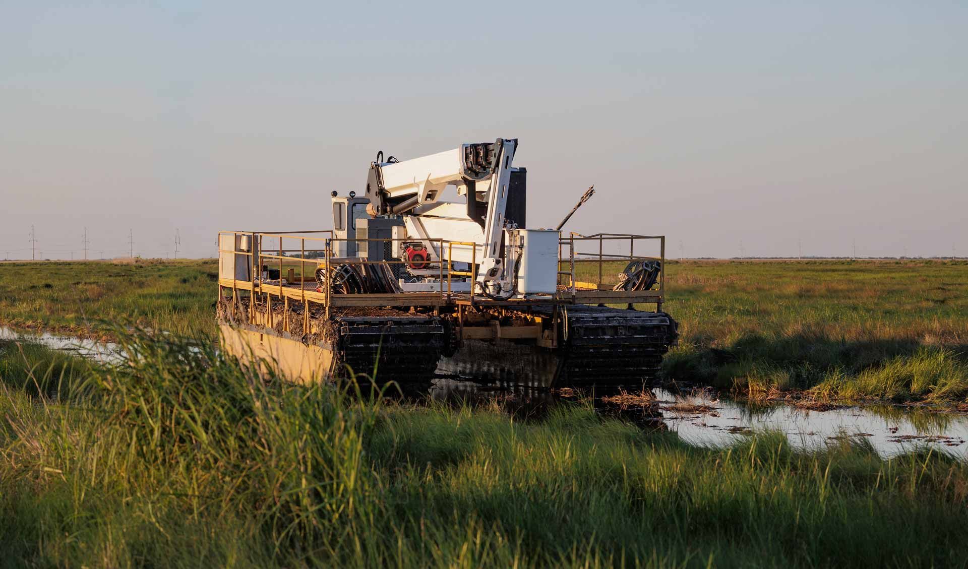 Bulldozer crossing a muddy wetland under a clear sky, surrounded by tall green grass.