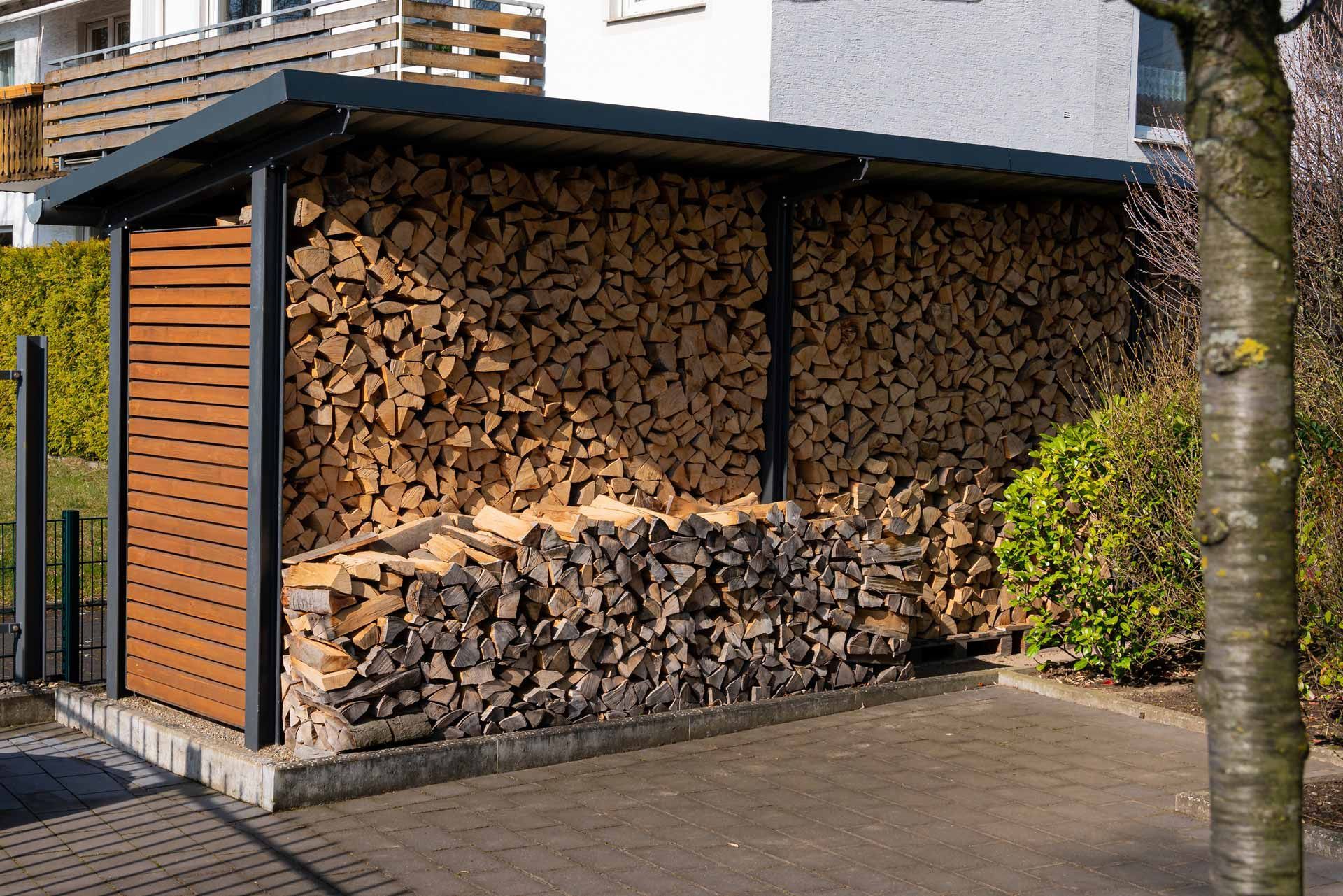 Wood shed stacked with cut firewood under a dark roof beside a paved driveway