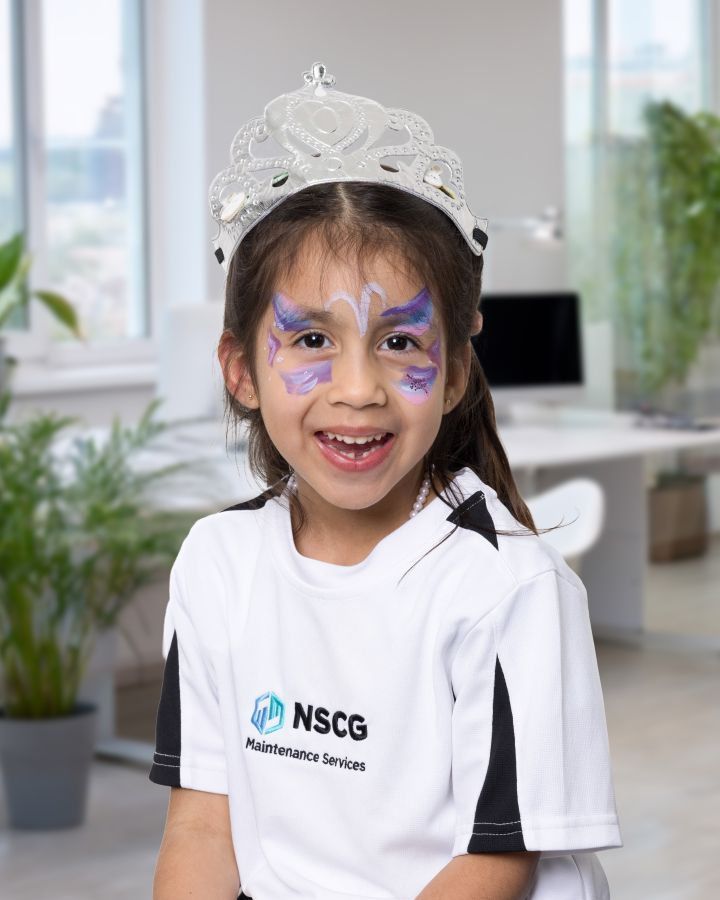 A smiling child wearing a silver tiara and purple butterfly face paint, dressed in a white NSCG branded t-shirt.
