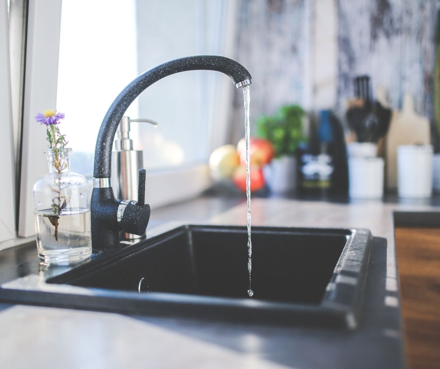 A close-up of a kitchen sink with a black faucet running water, a glass vase with a purple flower, and kitchen items.