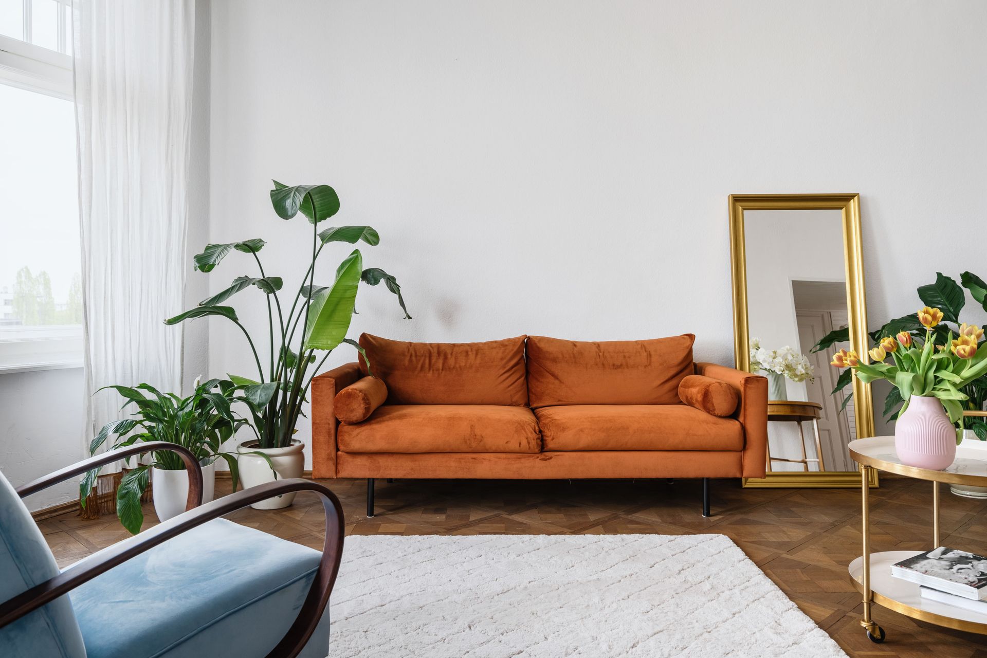 Orange velvet sofa in a white room with plants, a mirror, and a blue armchair.