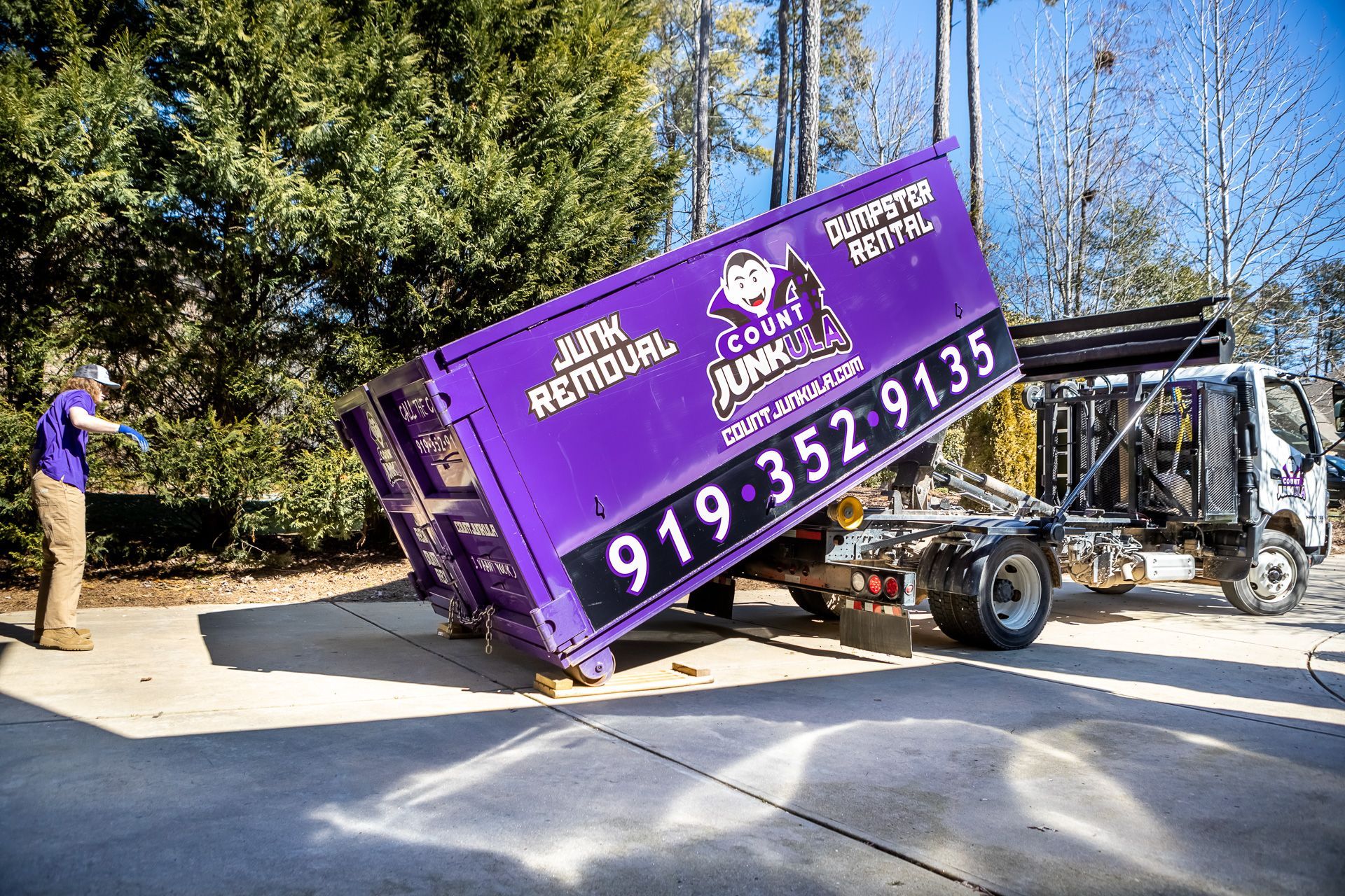 Purple dumpster being unloaded from a truck by a man in a yard, phone number on container.