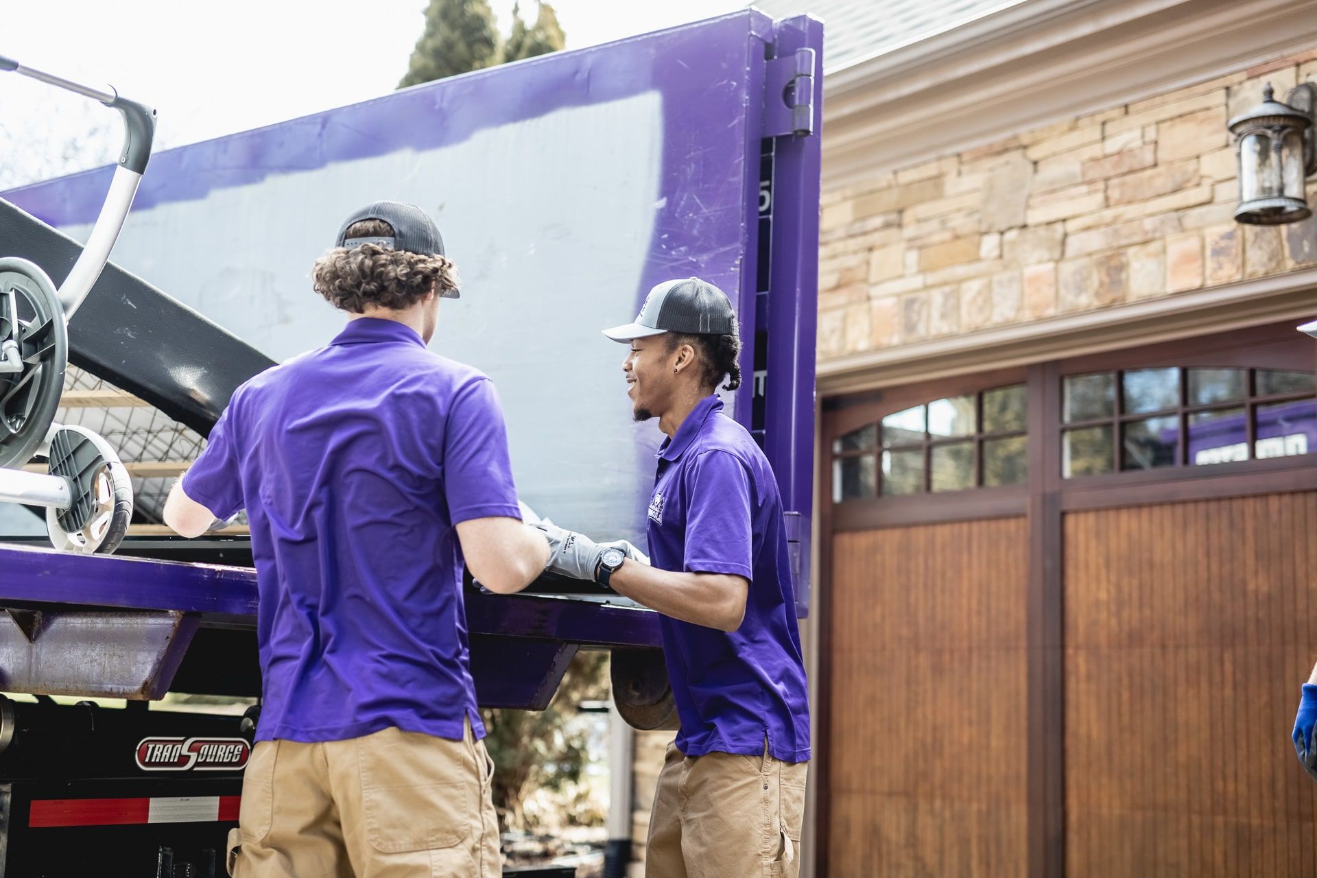 Two people in purple shirts load a large item onto a purple truck near a garage.