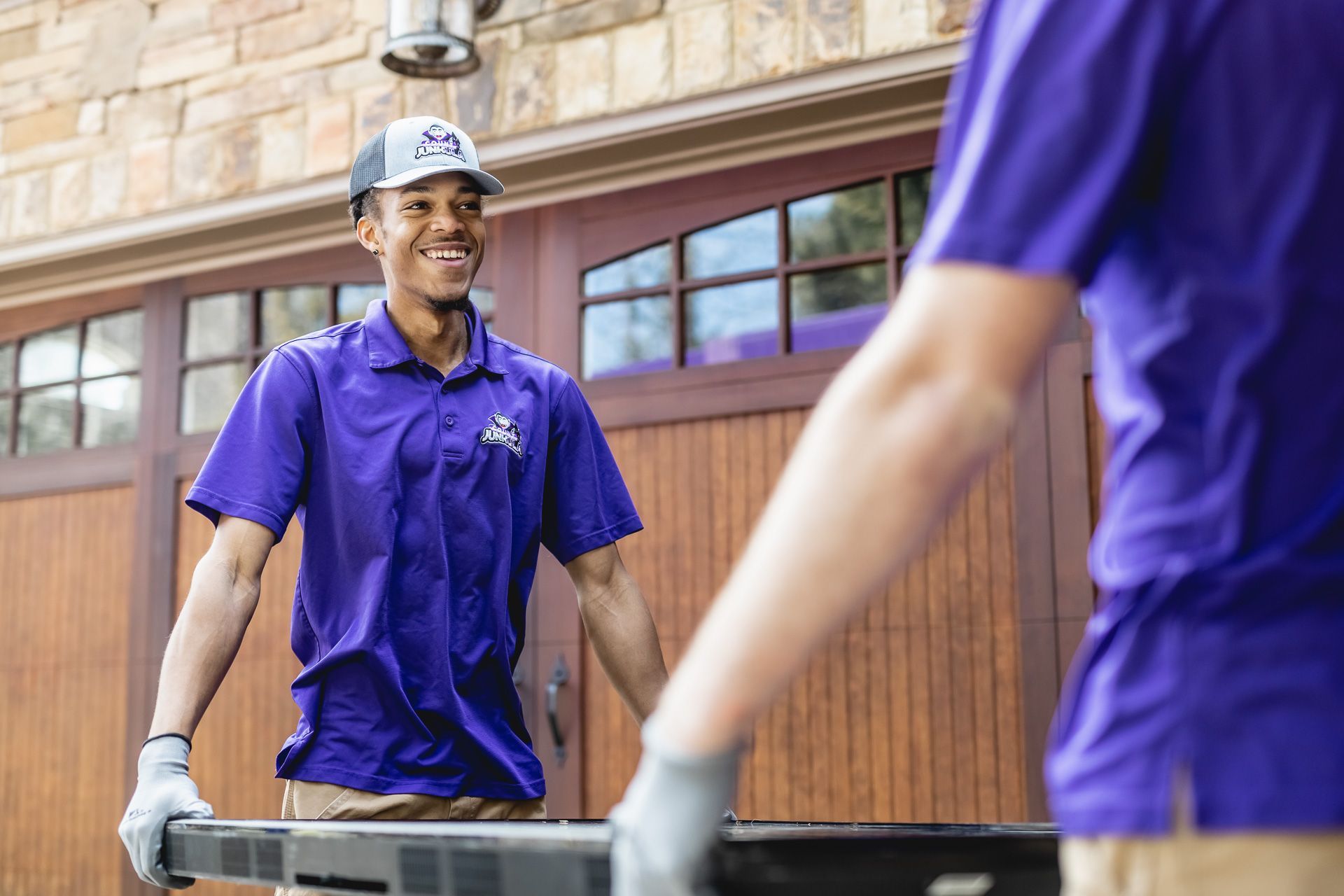 Two men in purple shirts and gloves carrying a black object; one smiles outside a garage.