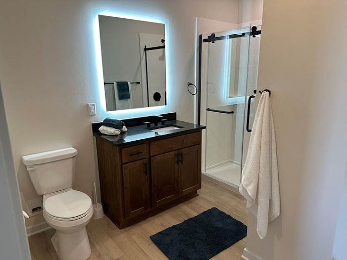 Bathroom with a lighted mirror above a dark wood vanity, toilet, and shower with a sliding black door.