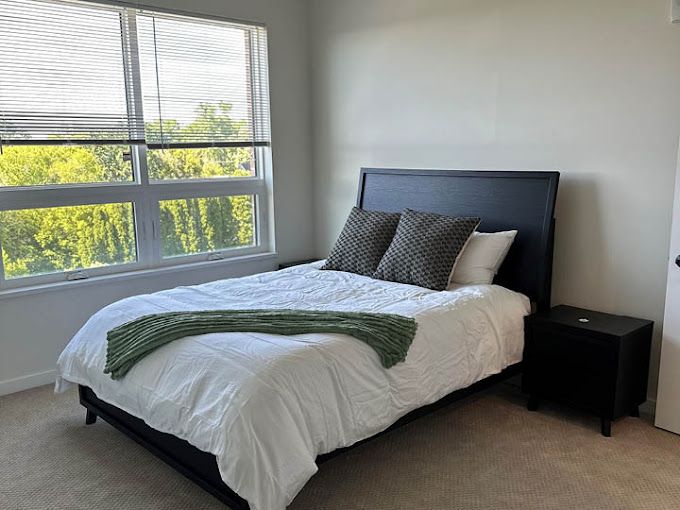 Bedroom with bed, nightstand, and window with blinds. White bedding, green throw, and neutral walls.