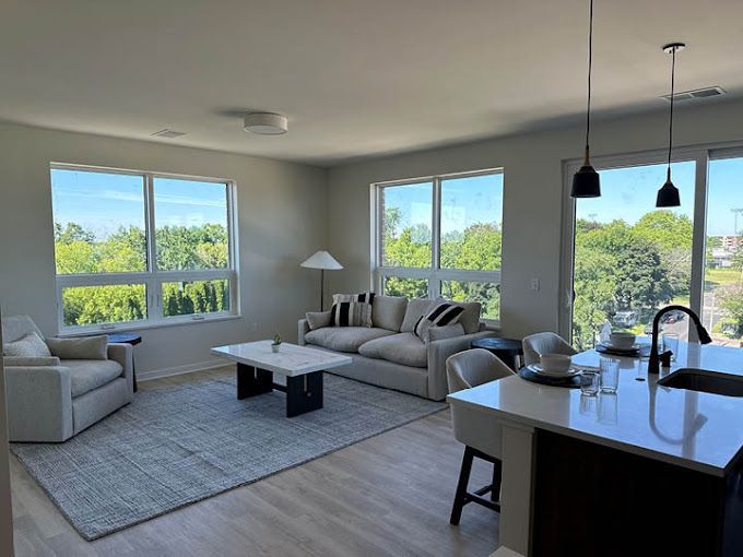 Living room with large windows overlooking trees. Includes couch, armchair, rug, and kitchen island with hanging lights.