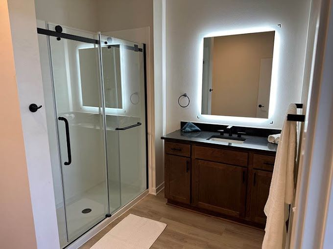 Bathroom with glass shower, lit mirror, dark cabinets, and light flooring.
