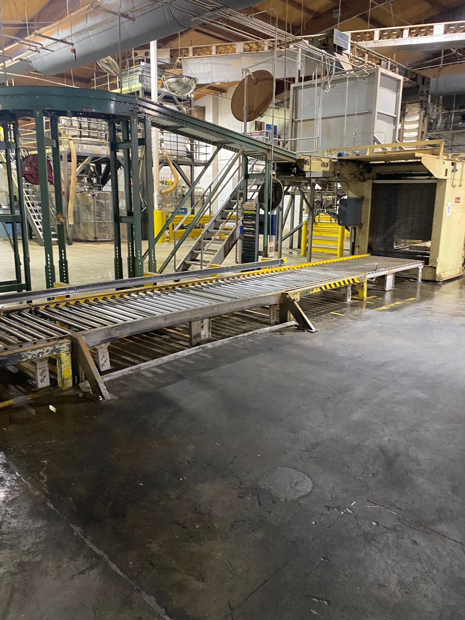 Industrial factory interior with metal framework, yellow railings, and a conveyor line over a wet floor.