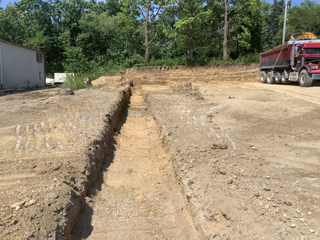Construction site with long trench, truck, and building. Earth and trees in the background. Sunny day.
