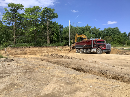 Excavator loading dirt into a red dump truck at a construction site with trees and a blue sky.